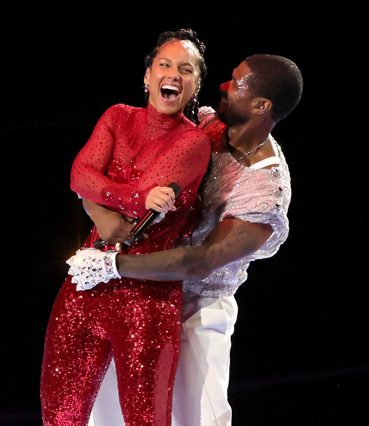 Alicia Keys laughs while holding a mic in a sparkly red catsuit while Usher in a white outfit holds his arm around her waist