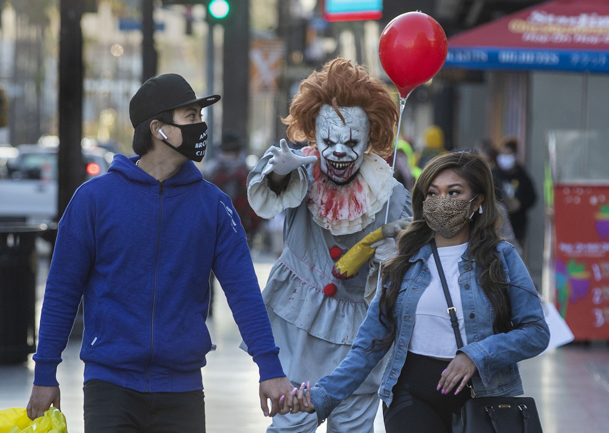A street performer dressed as Pennywise the clown holds an arm out toward a masked couple walking on the sidewalk