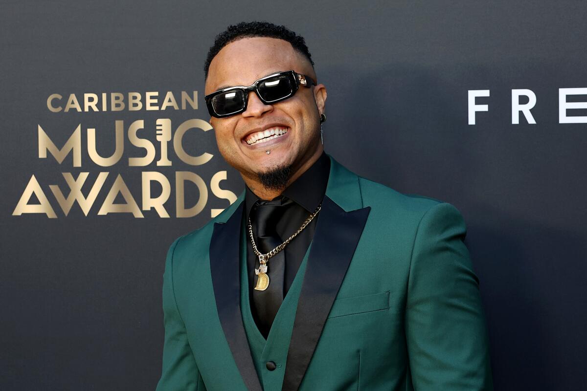Ricardo Drue flashes a big smile as he poses in a dark green and black three-piece suit and dark sunglasses