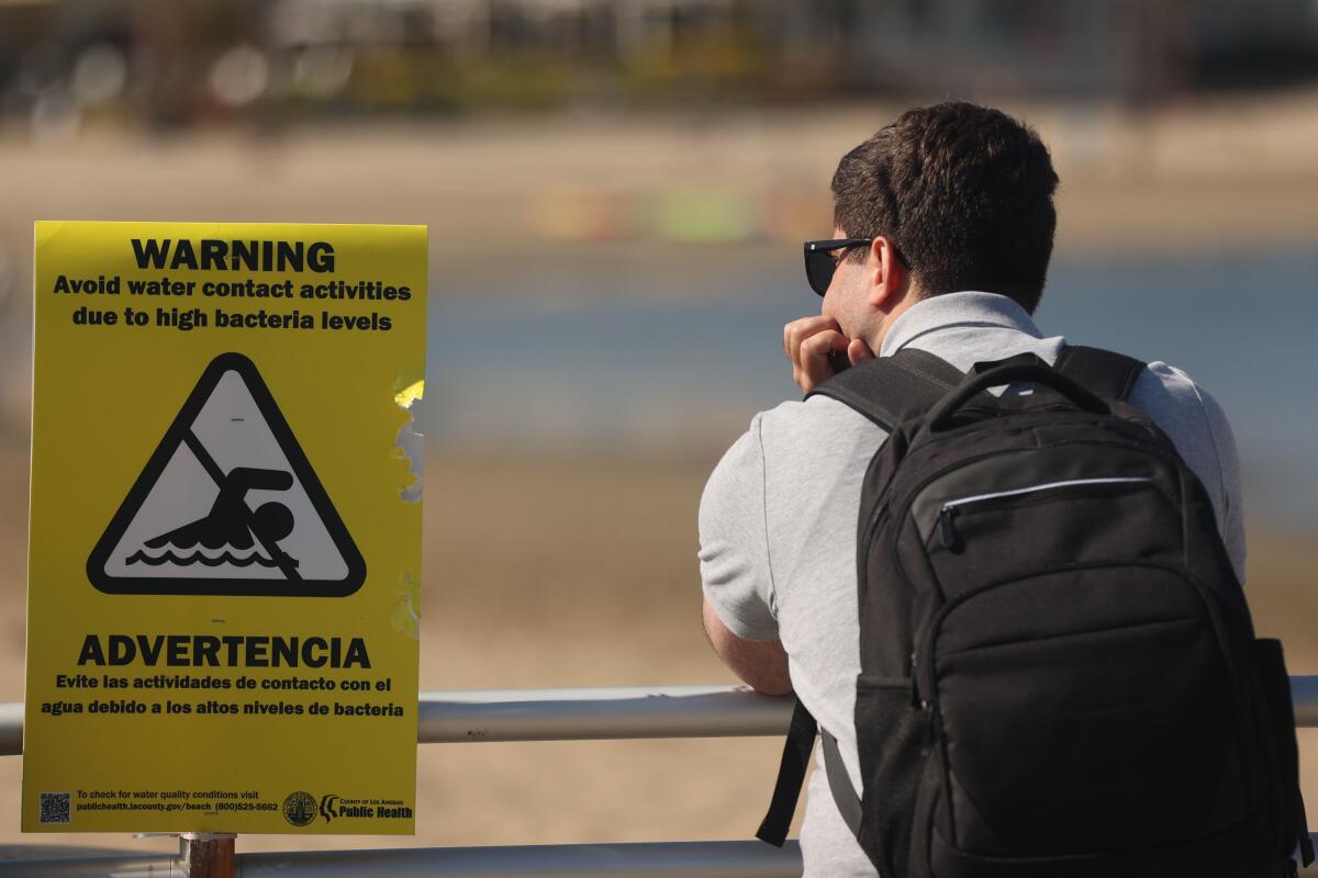 A man leans on a railing at a beach near a sign reading "Warning: Avoid water contact activities due to high bacteria levels"