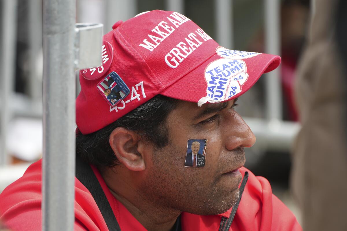 Seorang pria di topi Maga dan stiker wajah Trump di pipinya tampak di sebelah kanan.