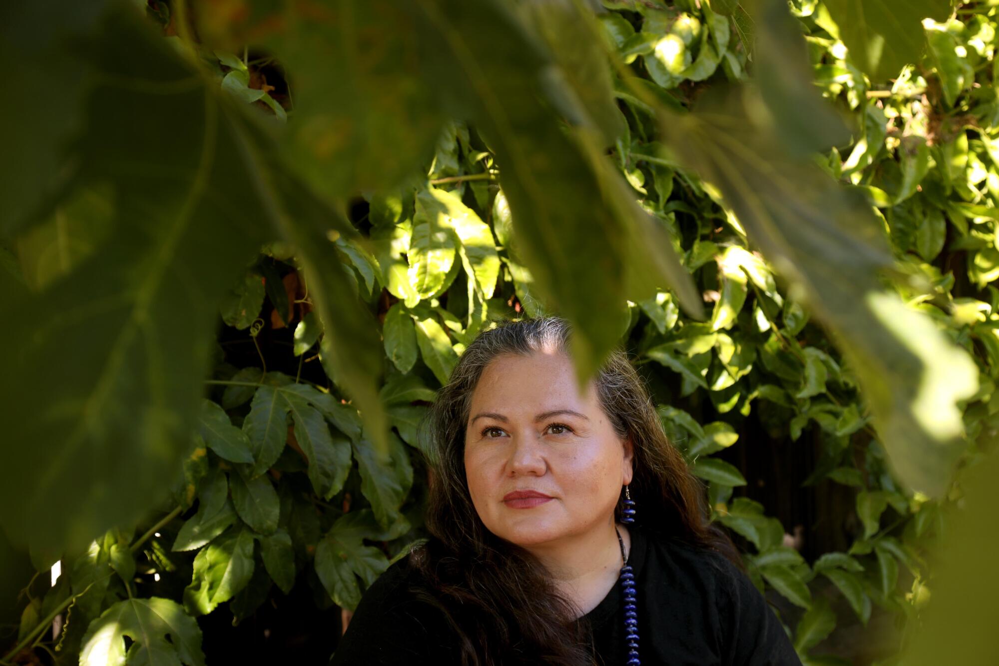 A woman with dark hair, in dark clothes, is surrounded by green plants 
