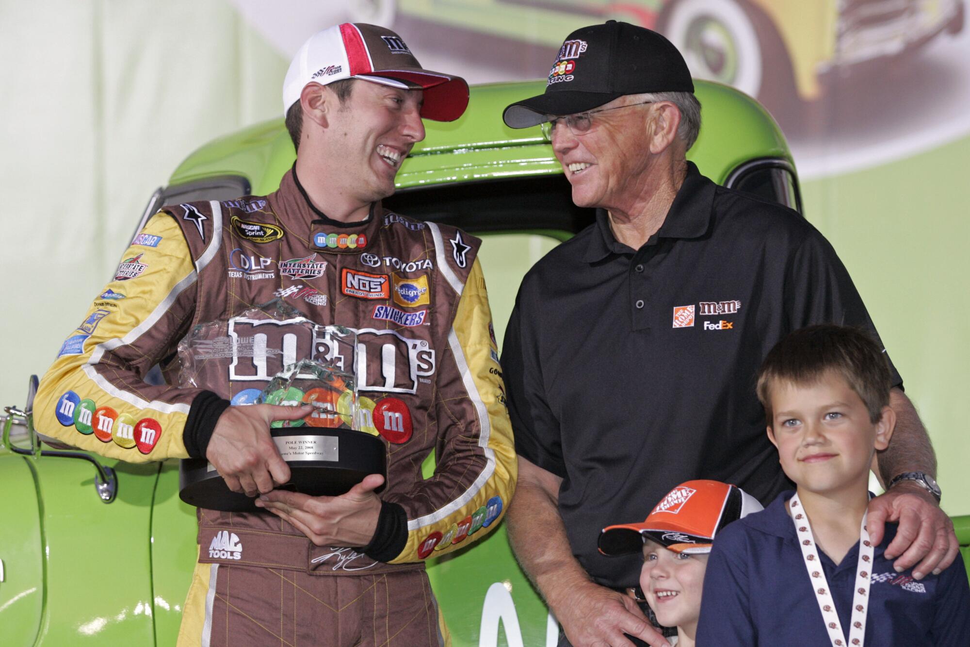 NASCAR driver Kyle Busch holds a trophy and jokes with team owner Joe Gibbs, who is behind two boys.