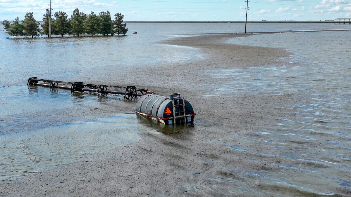A sheen on the surface of floodwaters surrounding half-submerged farm equipment and a barrel tank