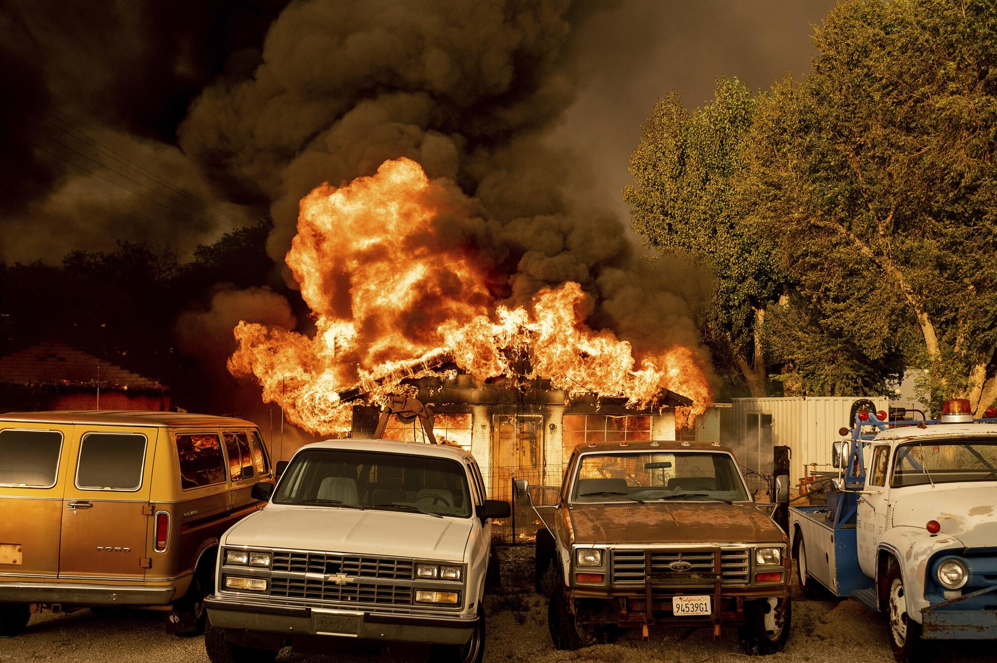 Flames destroy a home with three automobiles in the foreground