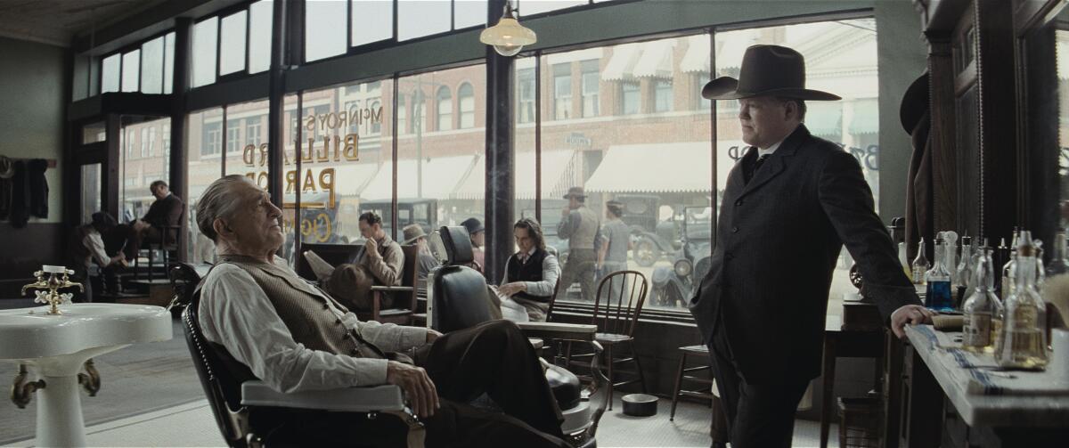 A man in a barber's chair speaks with a federal agent in a hat.