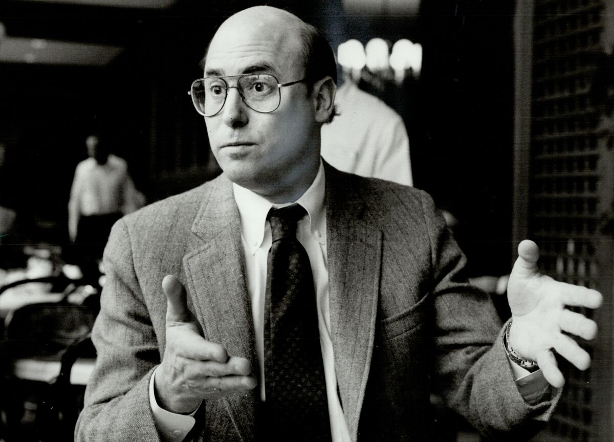 A black-and-white photo of a bald man with glasses in a suit and tie. He has his hands in an expressive position.
