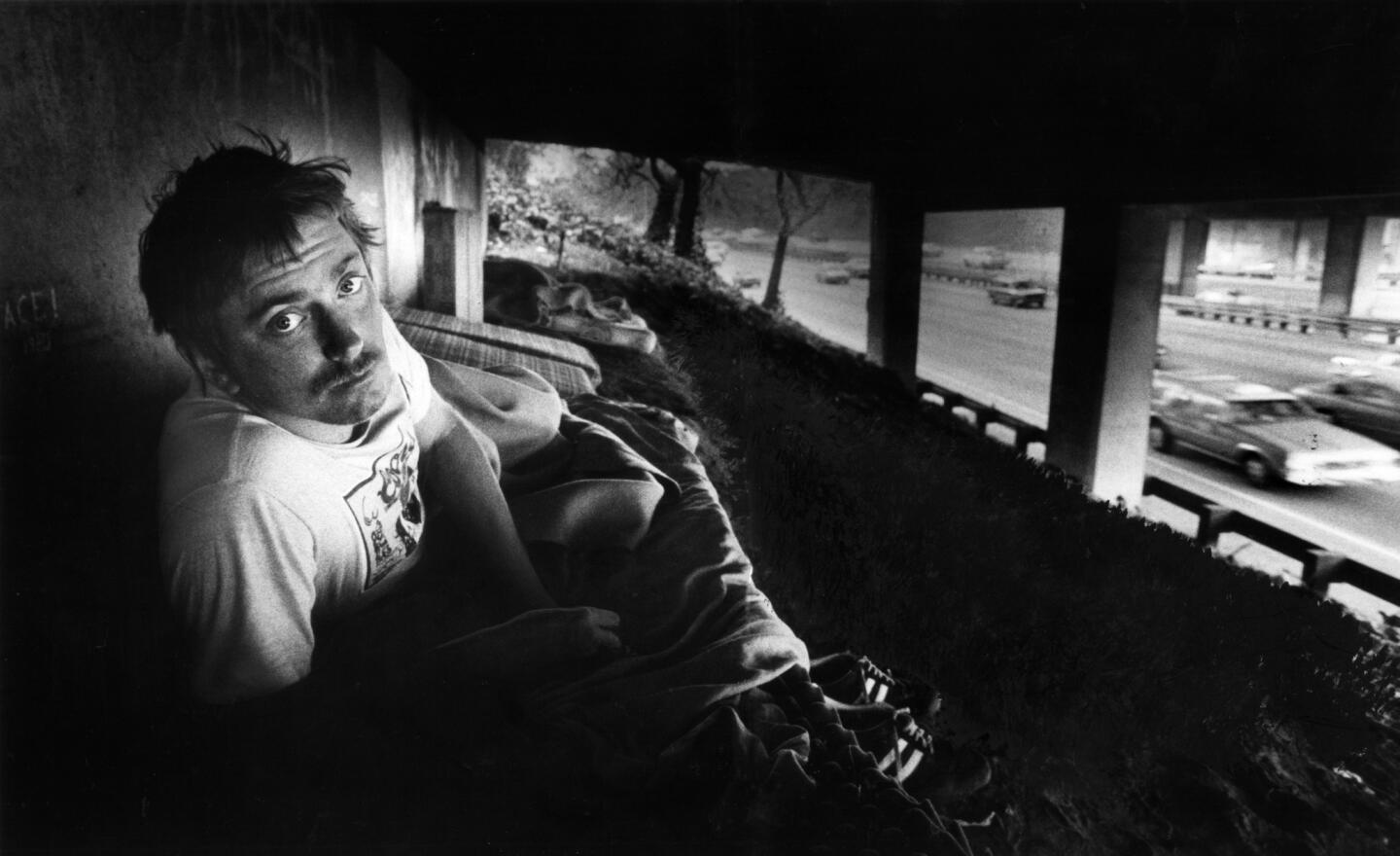 Nov. 8, 1982: Tom Kammer peers up from his bed, a mattress on dirt beneath an overpass of the 101 Freeway. Behind Kammer are other mattresses with one sleeping neighbor.