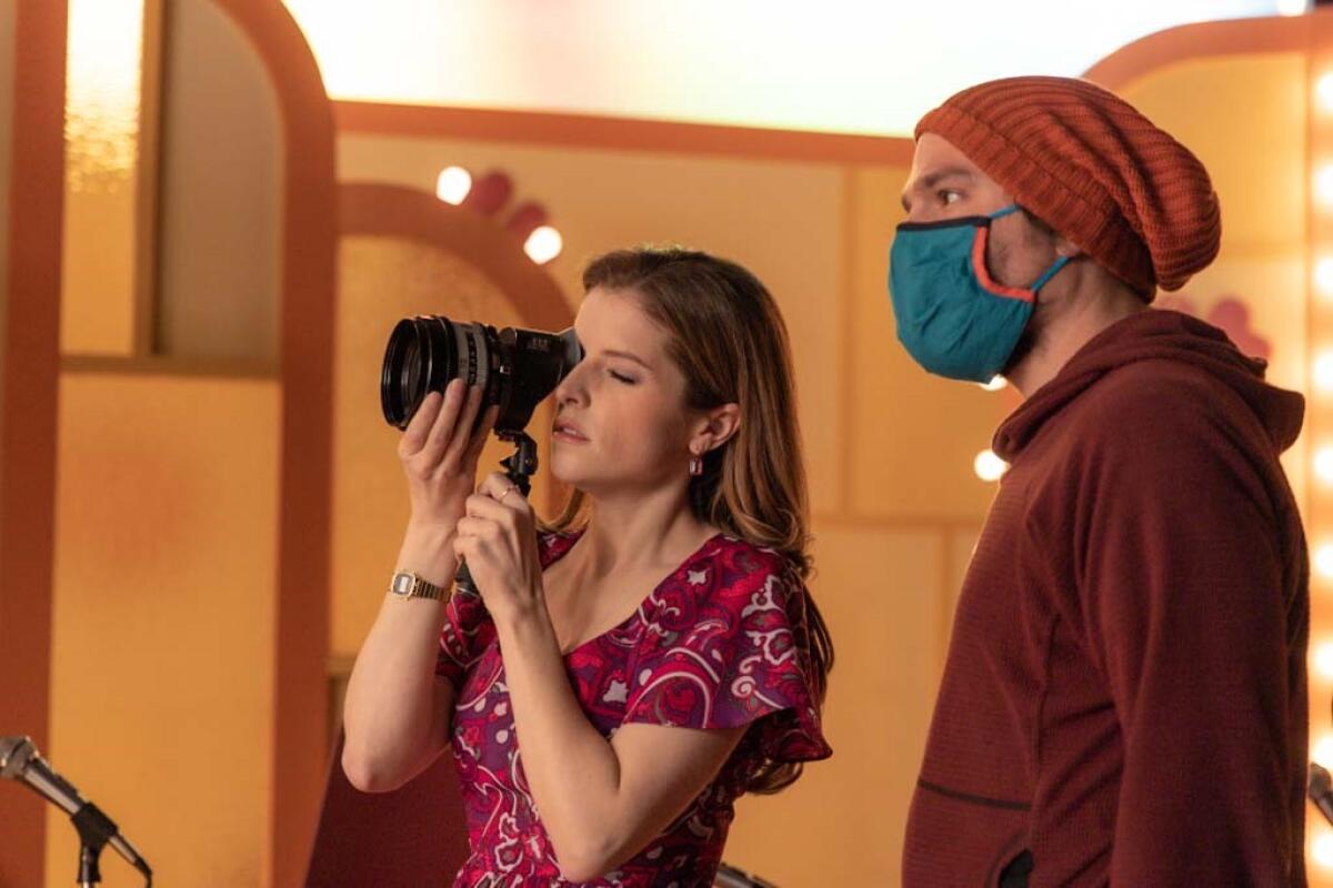 Anna Kendrick looks through a lens on a movie set, with a masked person standing next to her.