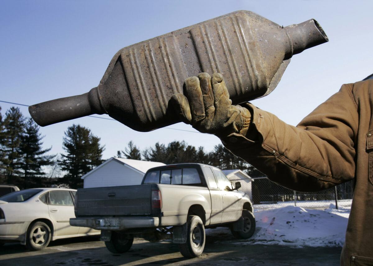 A catalytic converter is held up by a person. Two vehicles are also shown.