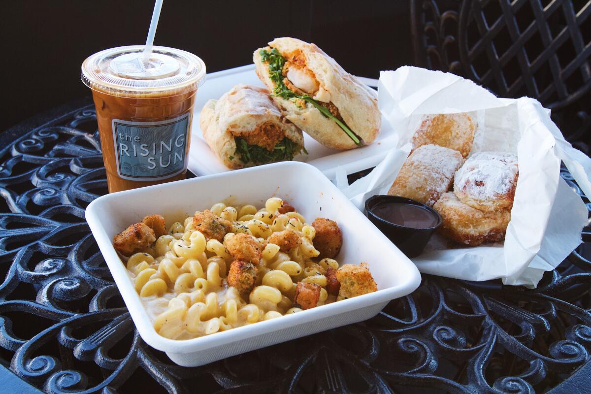 A horizontal photo of a shrimp po' boy, bag of beignets, fried-okra mac and cheese, and iced chicory coffee on a black table