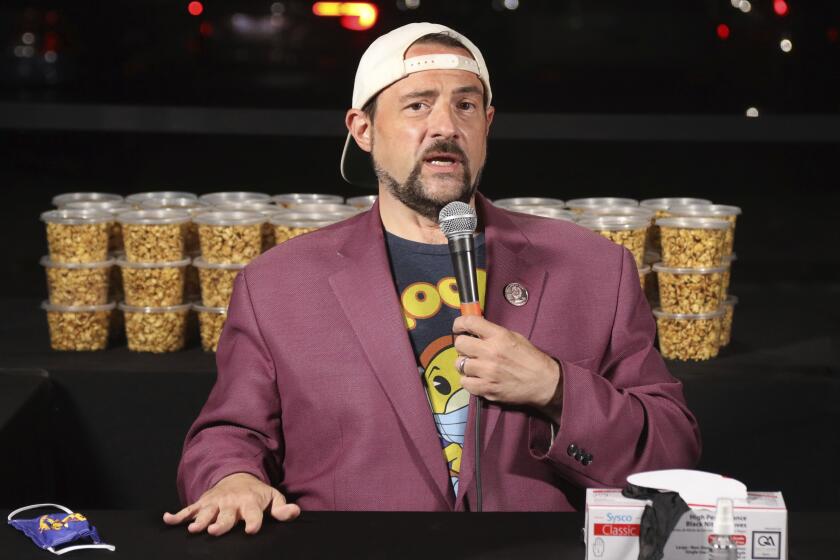 A man wearing a t-shirt, blazer and backwards cap sits at a table with popcorn behind him