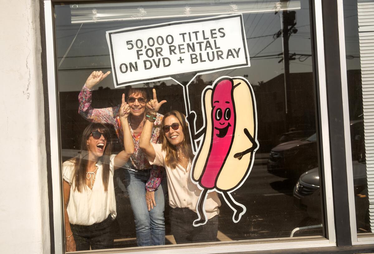 Three women in a video store window with a hot dog holding a sign