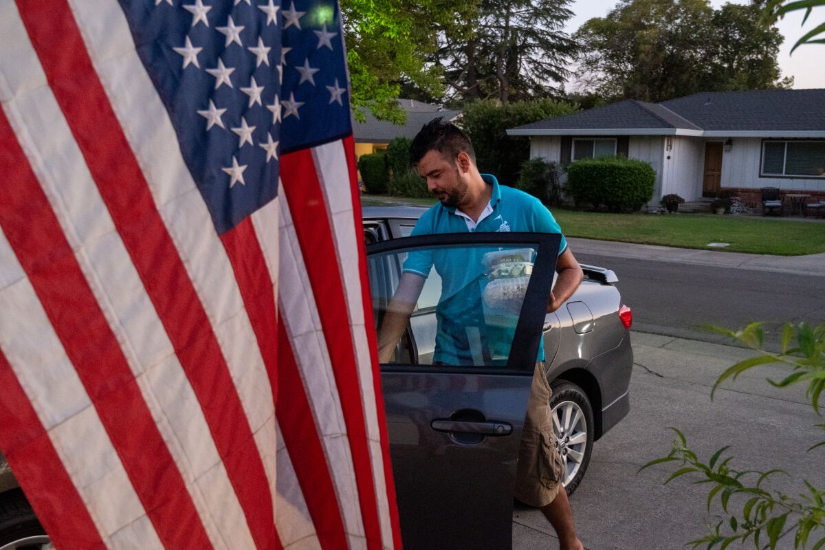 A bearded man in a blue polo shirt and shorts stands behind the open door of a car in a driveway. At left is a U.S. flag. 