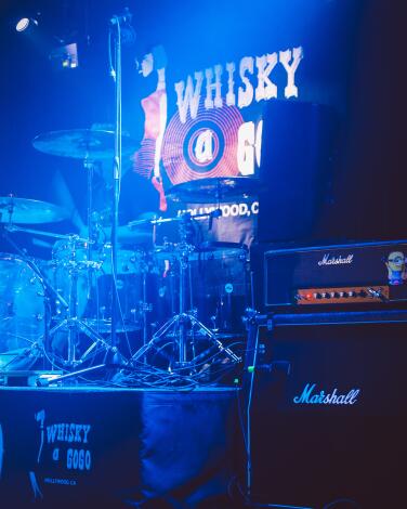 A drum kit and amplifiers in a blue-lighted room under a sign that says Whisky