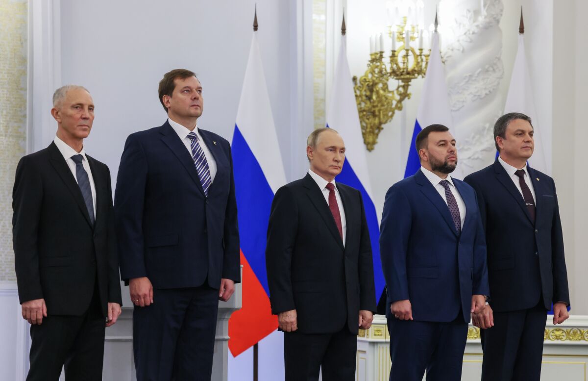 Five men with solemn expressions, in dark suits and ties, stand in front of white-blue-and-red flags
