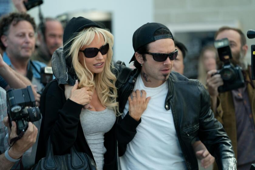 A woman and a man in dark glasses make their way through a throng of press
