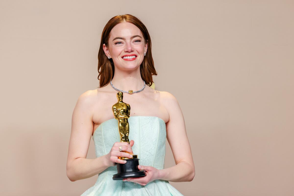 Emma Stone smiles in a pastel green strapless gown while holding an Oscar statuette with both hands