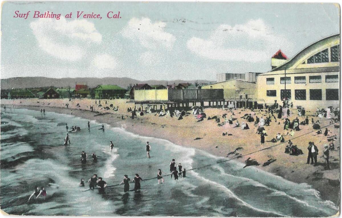 People play in the surf and sand. "Surf Bathing at Venice, Cal." the text on the front of the postcard reads. 