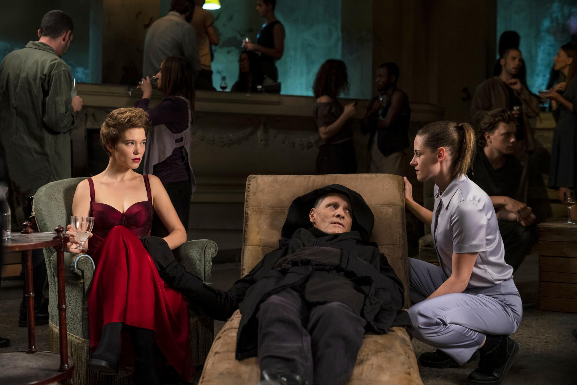 A woman in a red gown sits beside a man in black reclining and another woman crouches beside him in "Crimes of the Future."