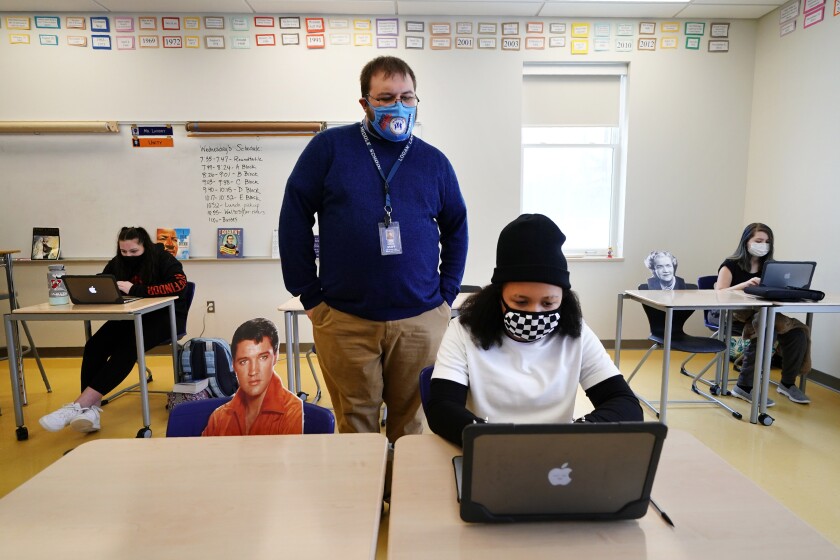 Social studies teacher Logan Landry looks over the shoulder of seventh grader Simone Moore as she works on a project while seated next to a cutout of Elvis Presley at the Bruce M. Whittier Middle School, Friday, Jan. 29, 2021, in Poland, Maine. With instruction time reduced as much as half by the coronavirus pandemic, many of the nation's middle school and high school teachers have given up on covering all the material normally included in their classes and instead are cutting lessons. Landry, put up cardboard cutouts to keep up social distancing, where instruction time has been cut in half by the hybrid model.(AP Photo/Robert F. Bukaty)