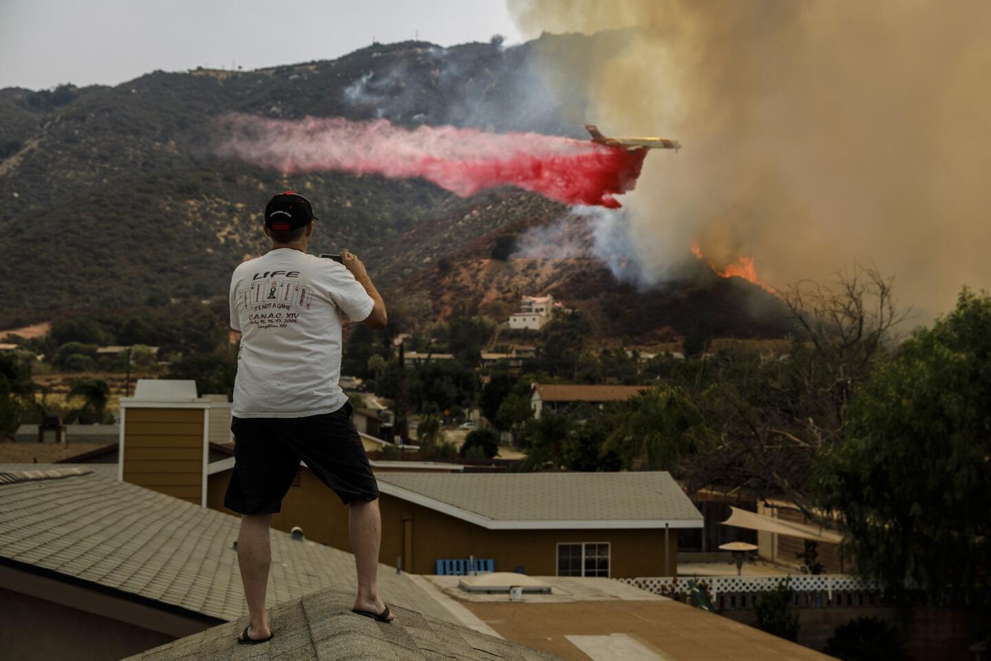 Jeff Qualls stands on his roof to watch an air tanker drop fire retardant on flames from the Holy fire as it make its way down the hillside in Lake Elsinore, Calif.