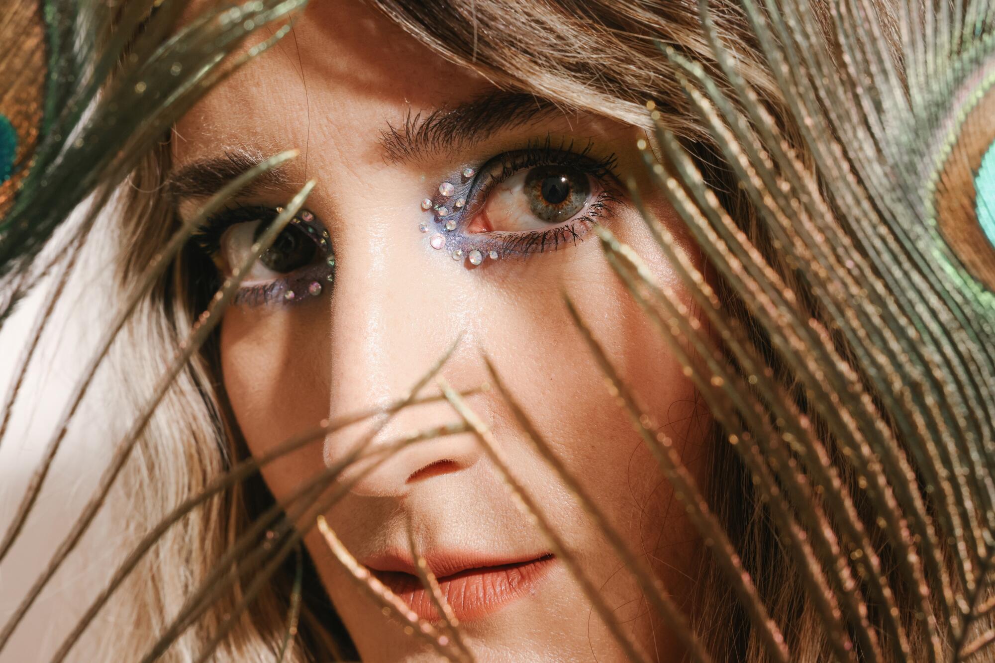 Closeup of a woman peeking between peacock feathers, her eyes adorned with eyeshadow and rhinestones. 