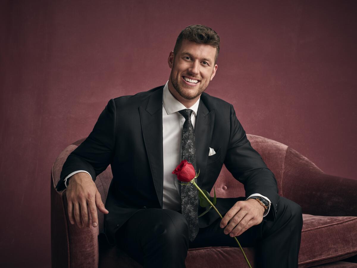 A man in a suit smiling, holding a rose, on a pink chair