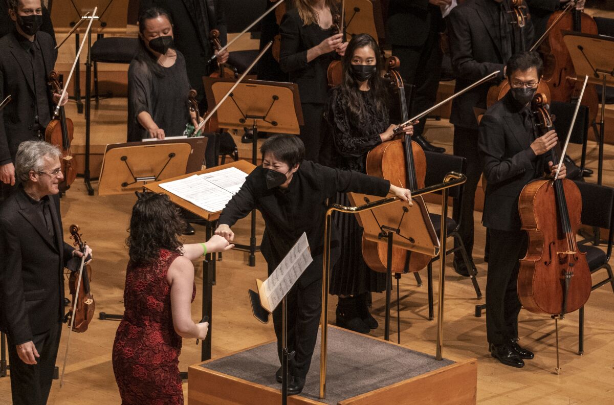 A conductor fist-bumps with a woman in a red dress as a man holding a violin looks on onstage at Disney Hall.