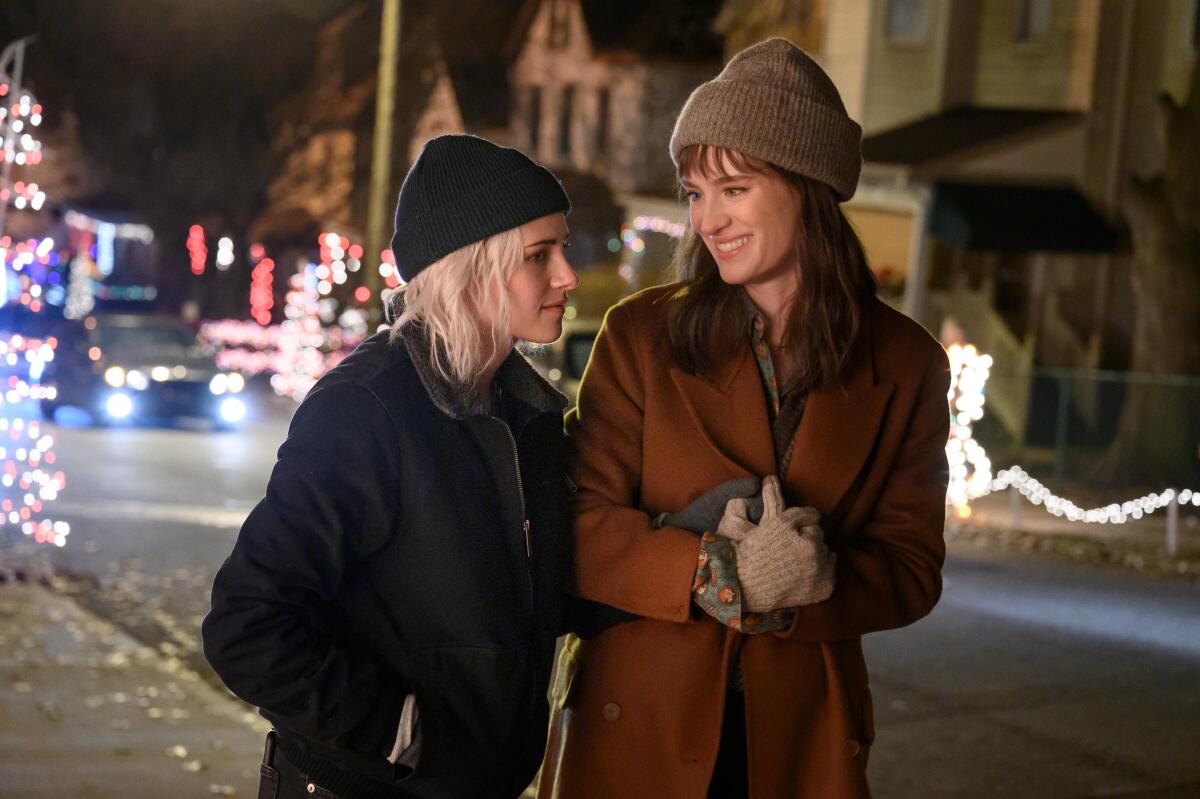 Kristen Stewart and Mackenzie Davis walk down a holiday-lighted street in the snow in a scene from "Happiest Season."