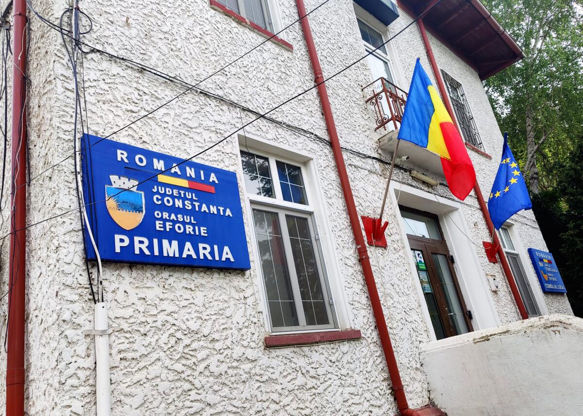 An exterior of a two-story white building with the Romanian flag and official plaques on its exterior. 