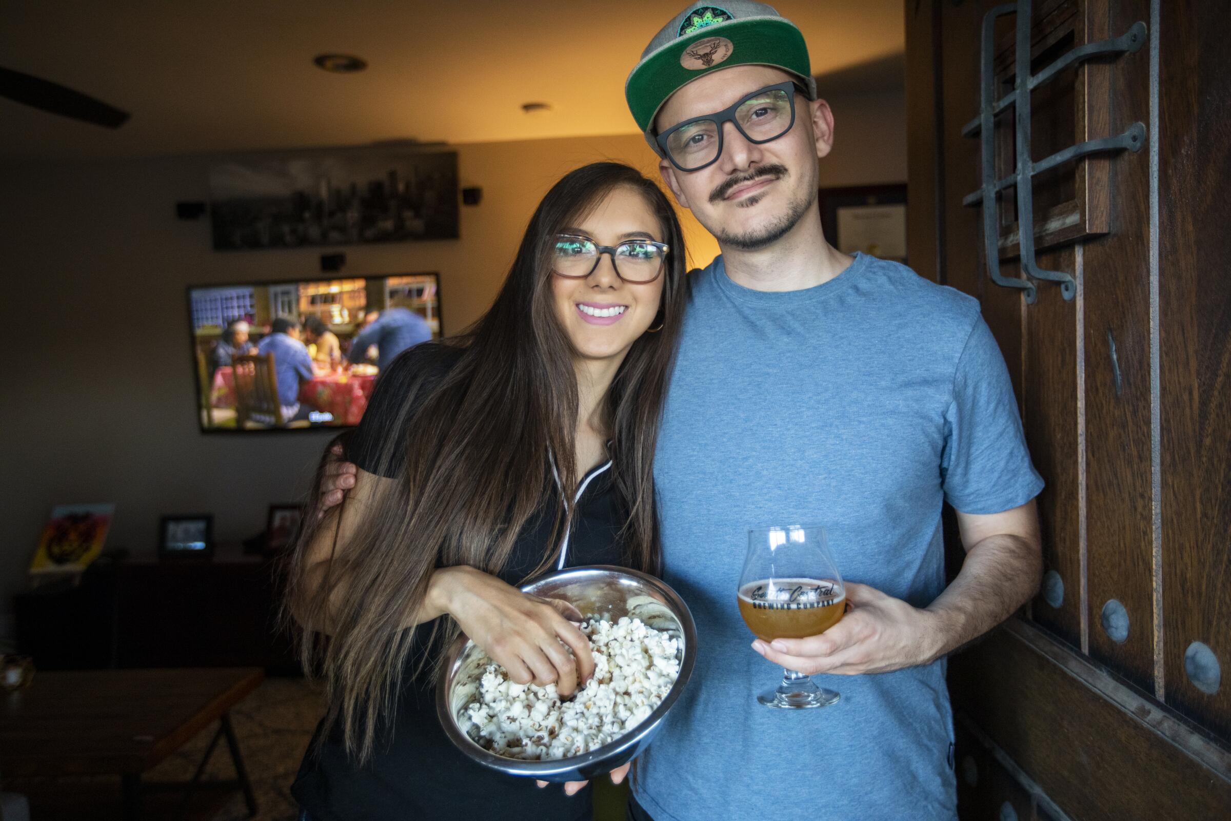 A woman with a bowl of popcorn and a man with a beer prepare to watch television.