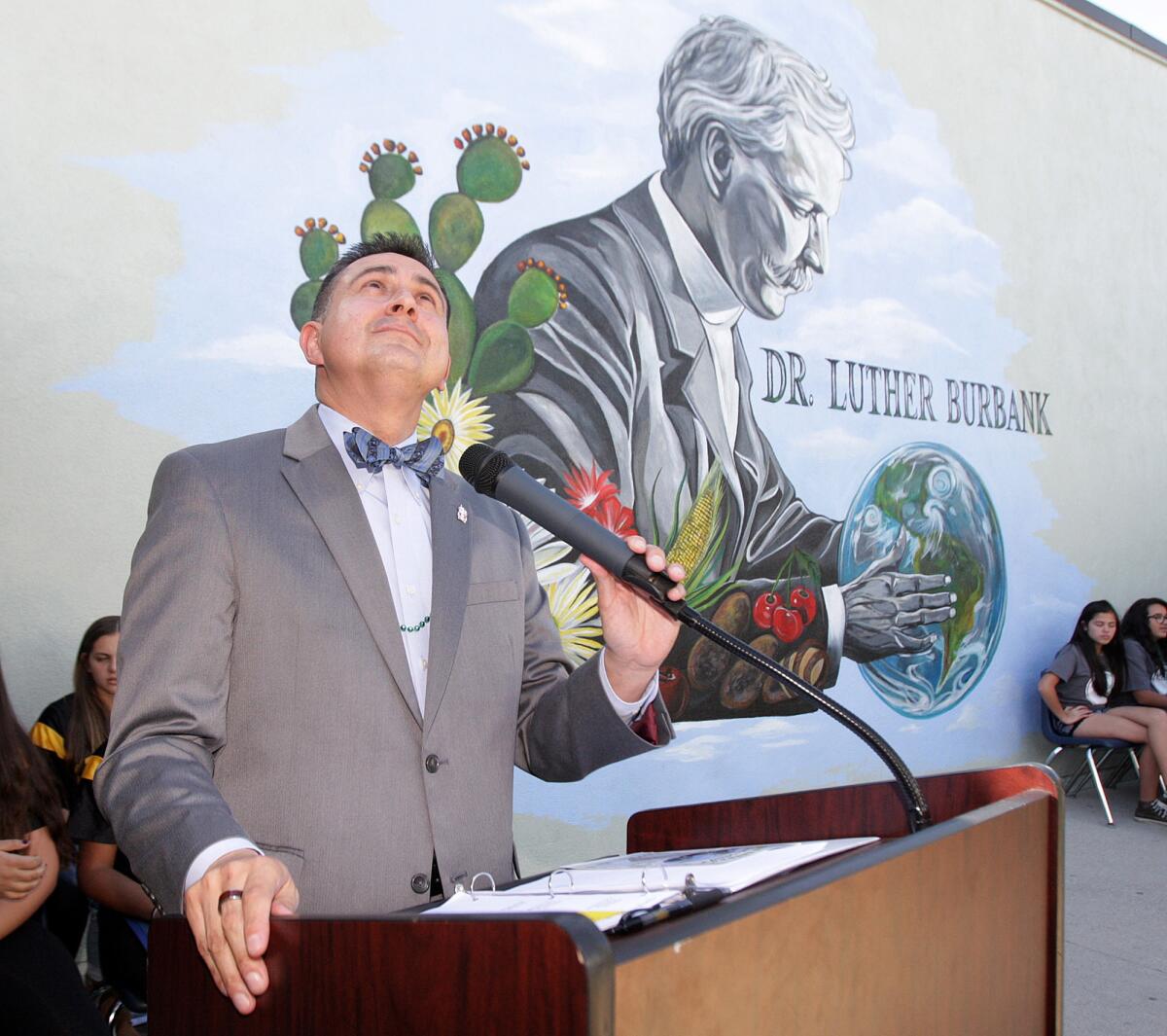 Luther Burbank Middle School Principal Oscar Macias takes the podium to close the ceremony as a jet, taking off from the Bob Hope Airport, flies overhead during the mural dedication and ribbon-cutting ceremony at the school on Tuesday, March 15, 2016. The mural depicts Dr. Luther Burbank and features about his life.