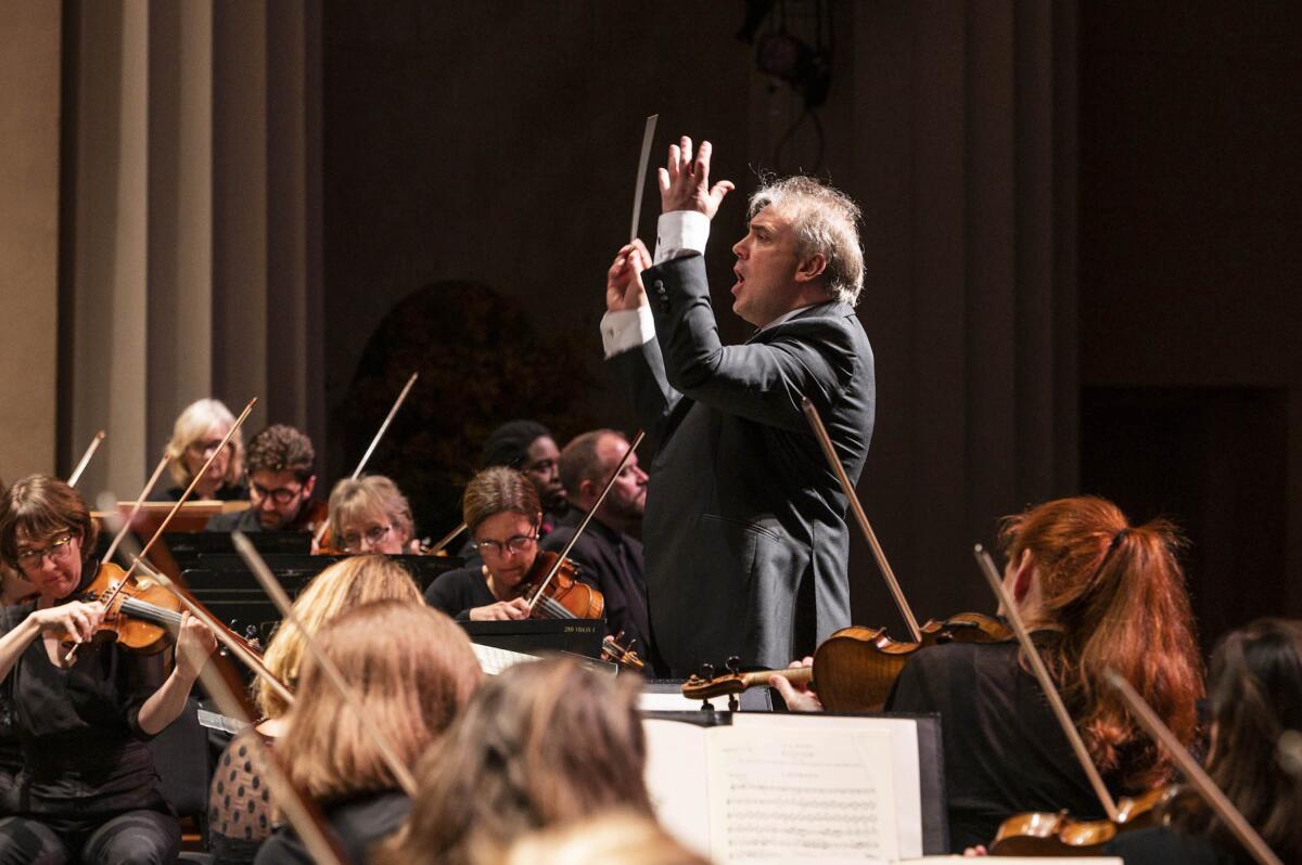 A man wearing a suit and holding a baton in his hand stands and conducts an orchestra.