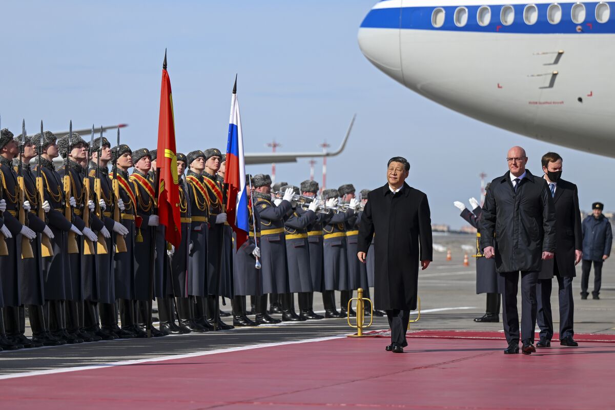 Ortada koyu renk paltolu bir adam, iki bayrağın yanında sıralanmış askeri üniformalı insanların yanından geçiyor.  Sağda bir uçak var 