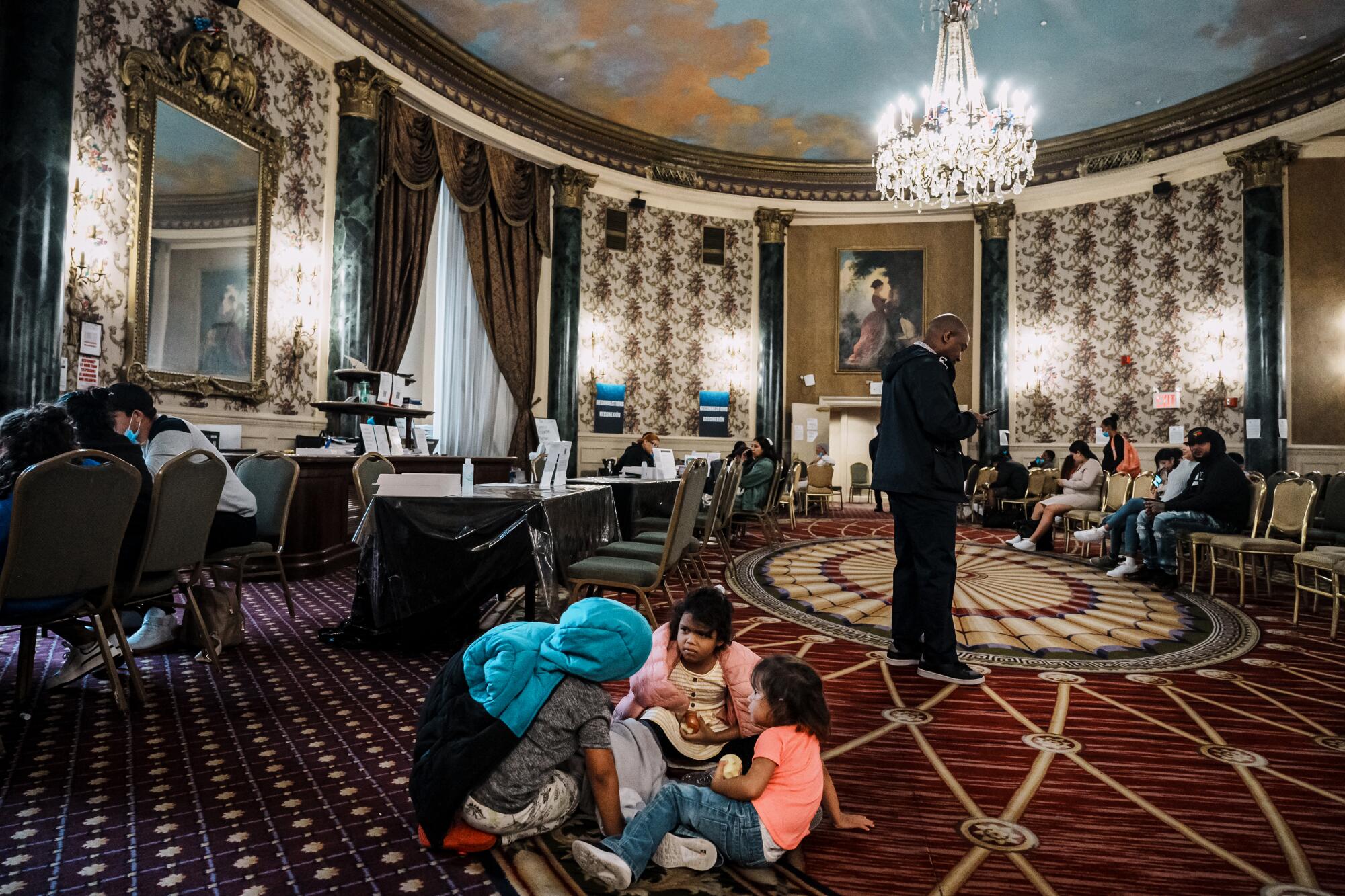 Children play on a carpeted floor while adults sit on chairs in an oval room.