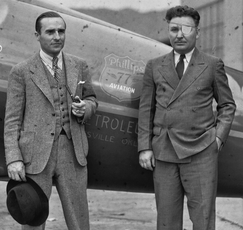 Two men in suits and ties stand outdoors next to a propeller plane. One man holds a fedora hat.