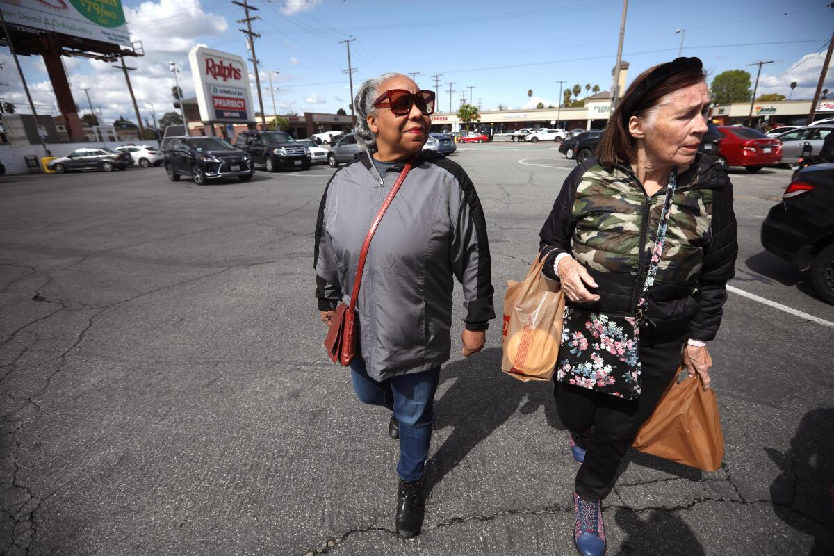 Two older women walk through a parking lot, one of them carrying grocery bags.