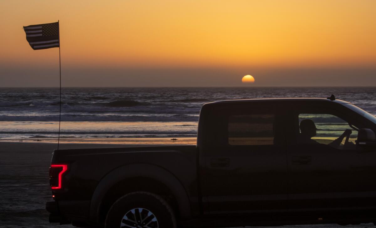 A pickup truck drives on the beach at sunset with a U.S. flag flying on the back
