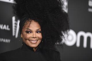 Janet Jackson wearing a black, high-neck top as she poses and smiles against a black background at a red carpet event