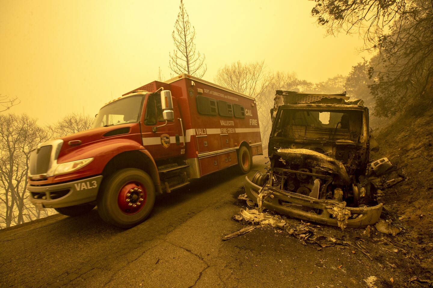 A red fire truck drives past the burned-out ruins of another vehicle on a road amid an orange, smoky haze.