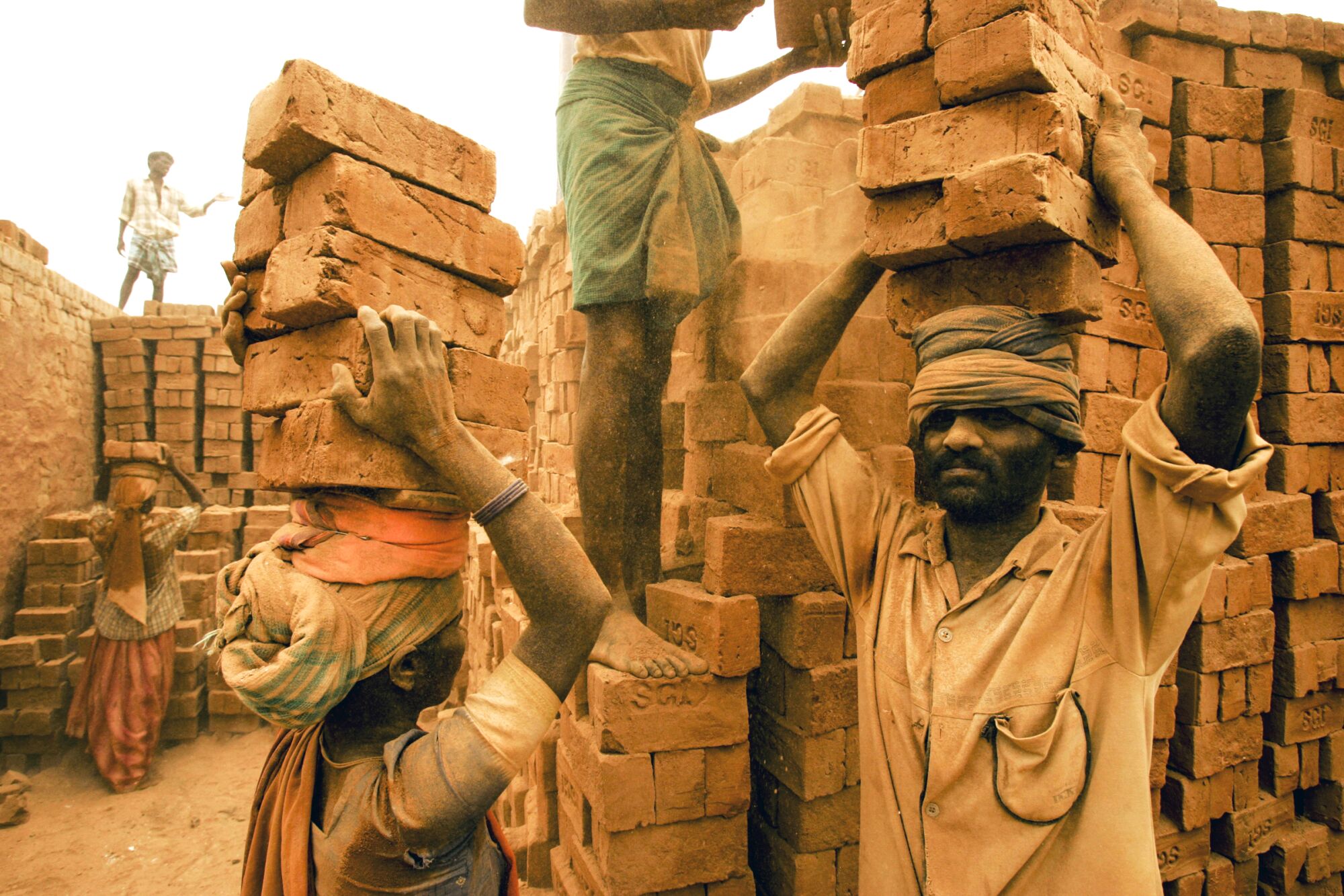 Migrant workers pile 20 bricks at a time on top of their heads, as dust falls in their faces