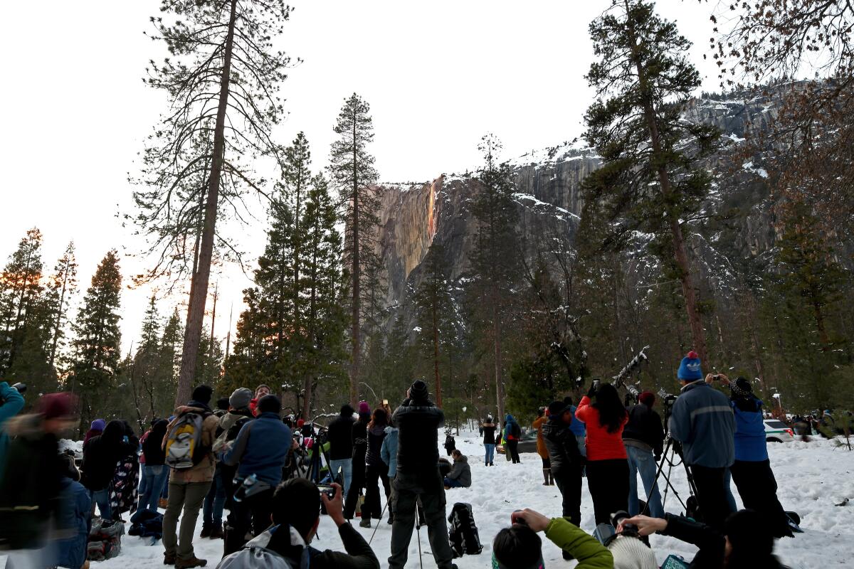 People gather at the base of a cliff to watch the light reflect off a waterfall.