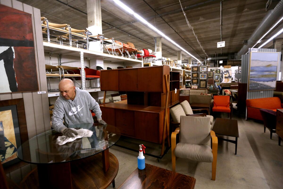 A man wipes down a round table in a large warehouse filled with furniture.