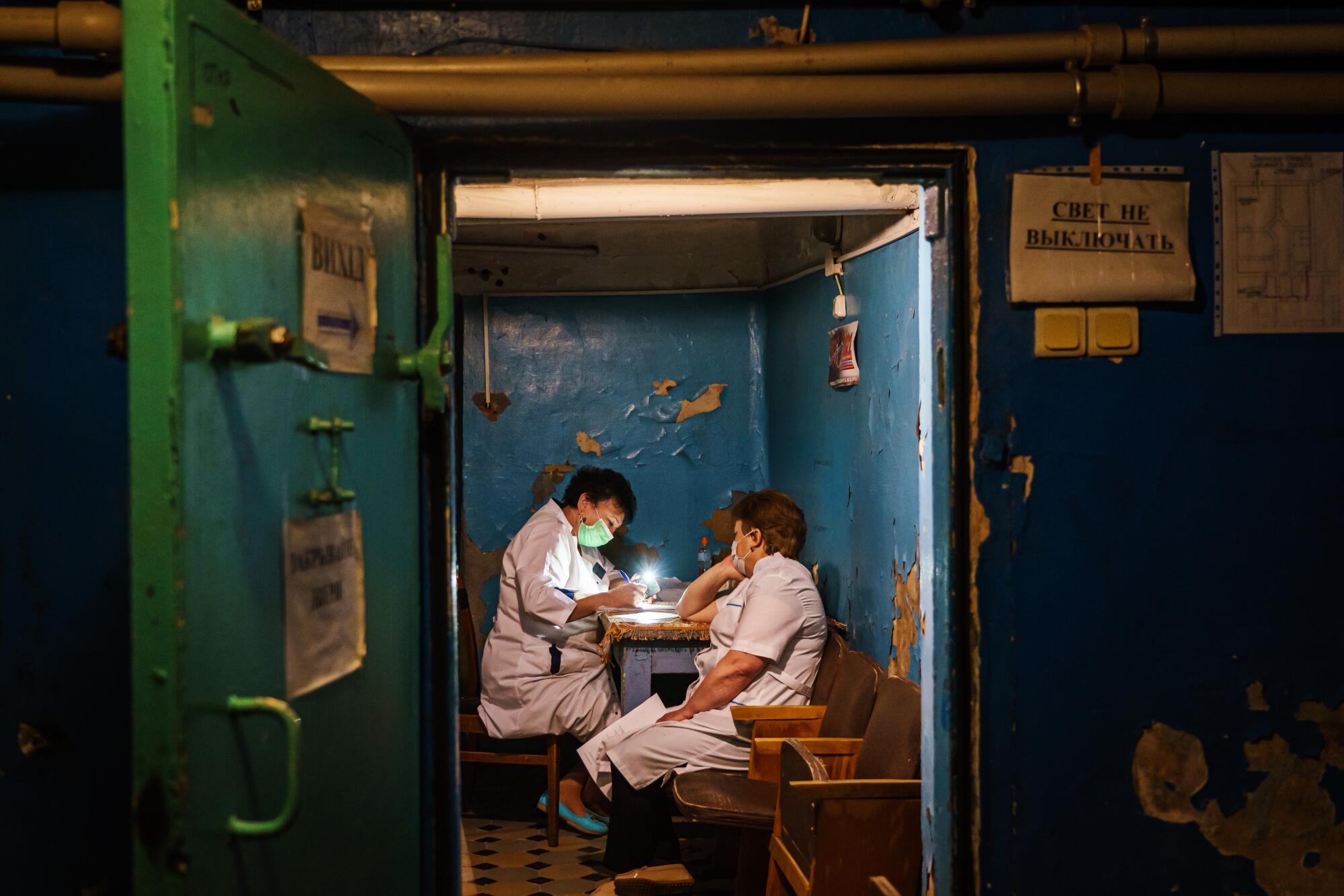 Two people in white hospital gowns sitting at a desk inside a dimly lit room in a dark underground shelter.