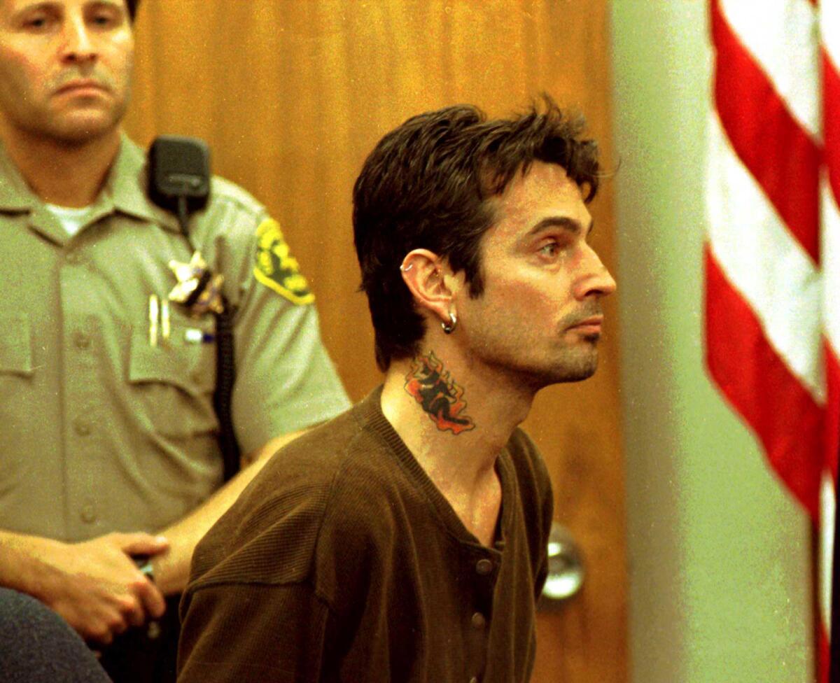 A man with short hair, earring and tattoo on his neck sits in court with a bailiff and U.S. flag in the background.