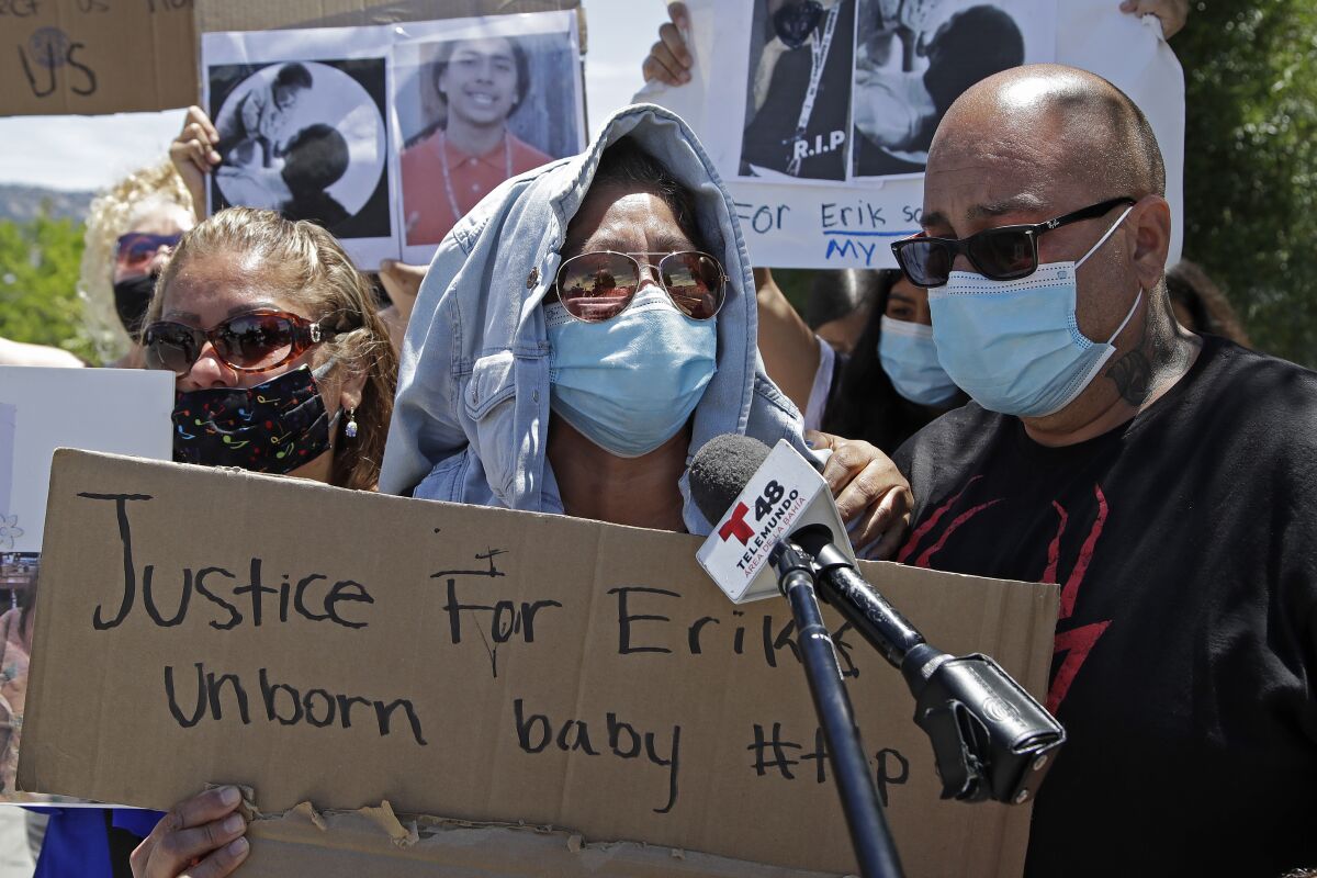 People wearing masks speak into a microphone and hold a cardboard sign that reads "Justice for Erik's unborn baby"