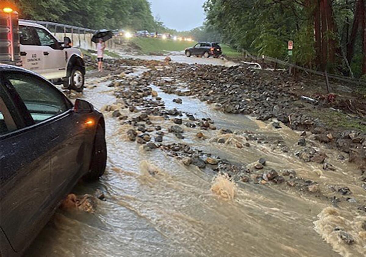 Vehicles come to a standstill near debris in a washed-out and flooded road.