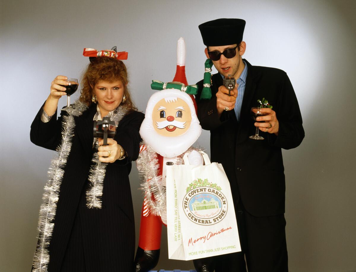 Kirsty MacColl and Shane MacGowan stand toasting with wine glasses, holding toy guns, next to an inflatable Santa.