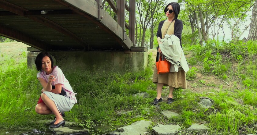 A woman crouches beneath a bridge over grass while another woman looks at her 